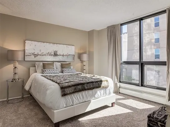 Bright staged bedroom with a white upholstered bed, neutral bedding, decorative pillows, abstract wall art, floor lamp, and large window with curtains letting in natural light.