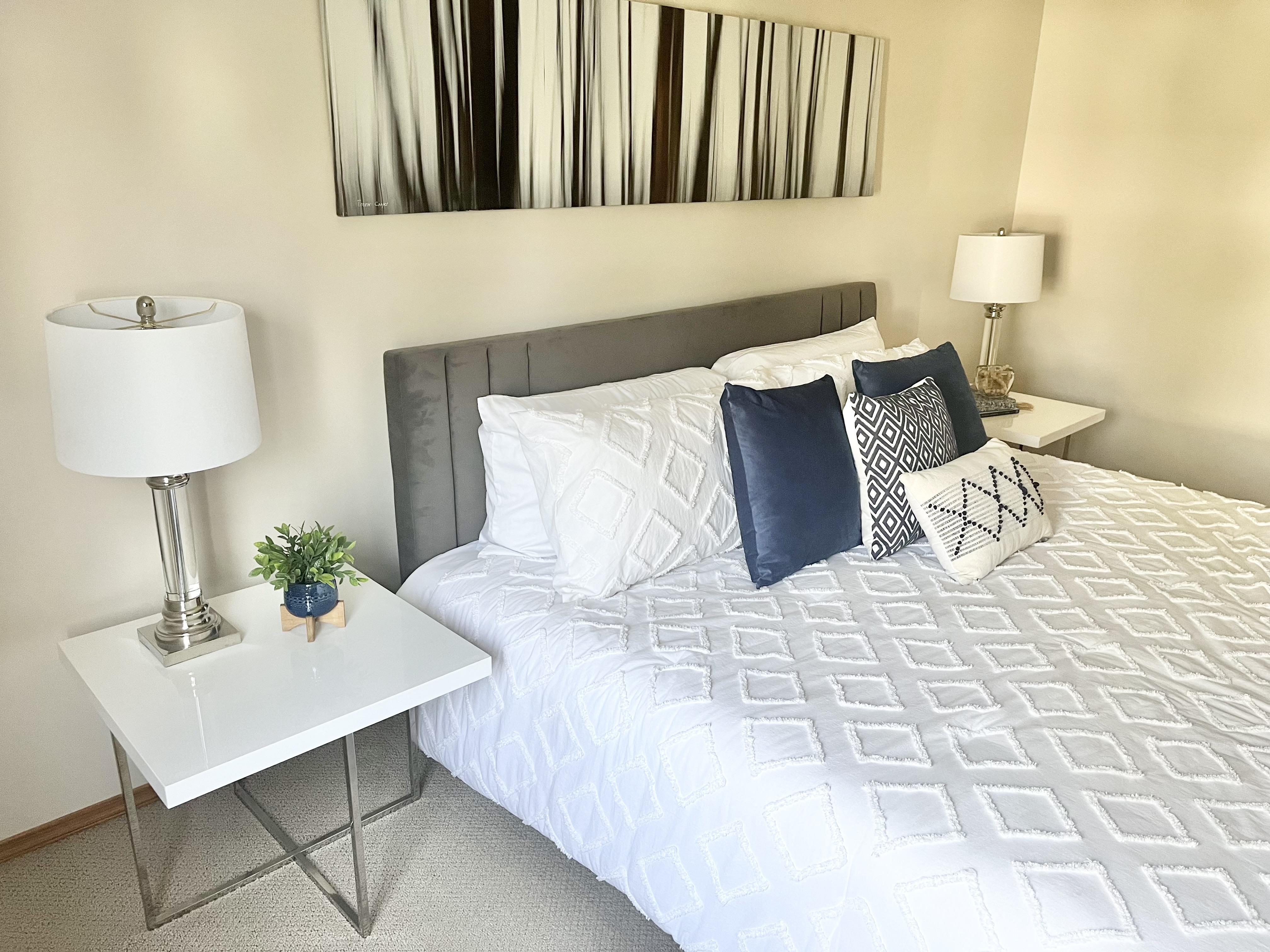 Staged bedroom with a gray upholstered headboard, white bedding with textured pattern, blue and patterned accent pillows, and modern bedside tables with lamps.