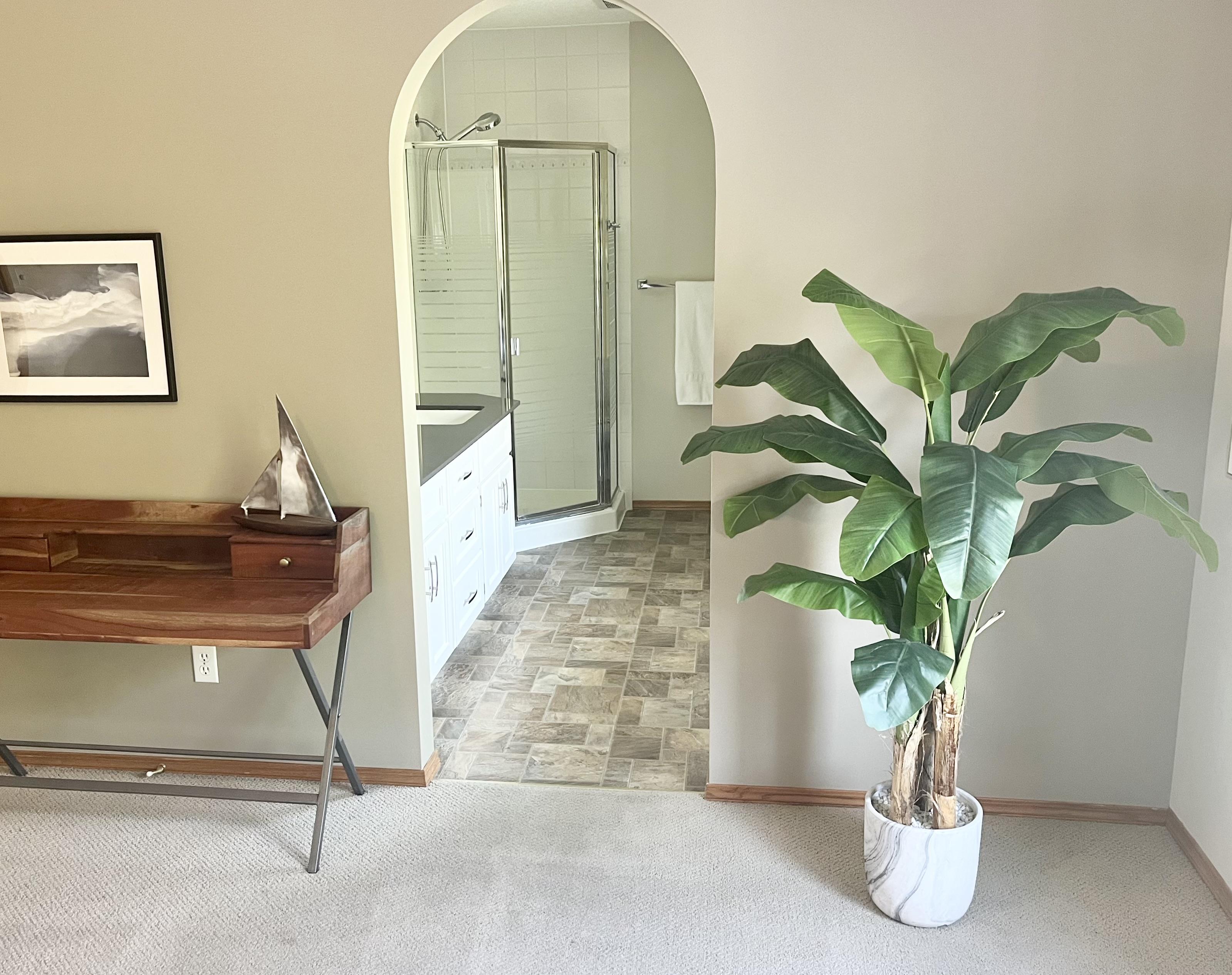 View through an arched doorway into a staged bathroom with a glass shower, vanity, and tile floor, with a large potted plant in the foreground.