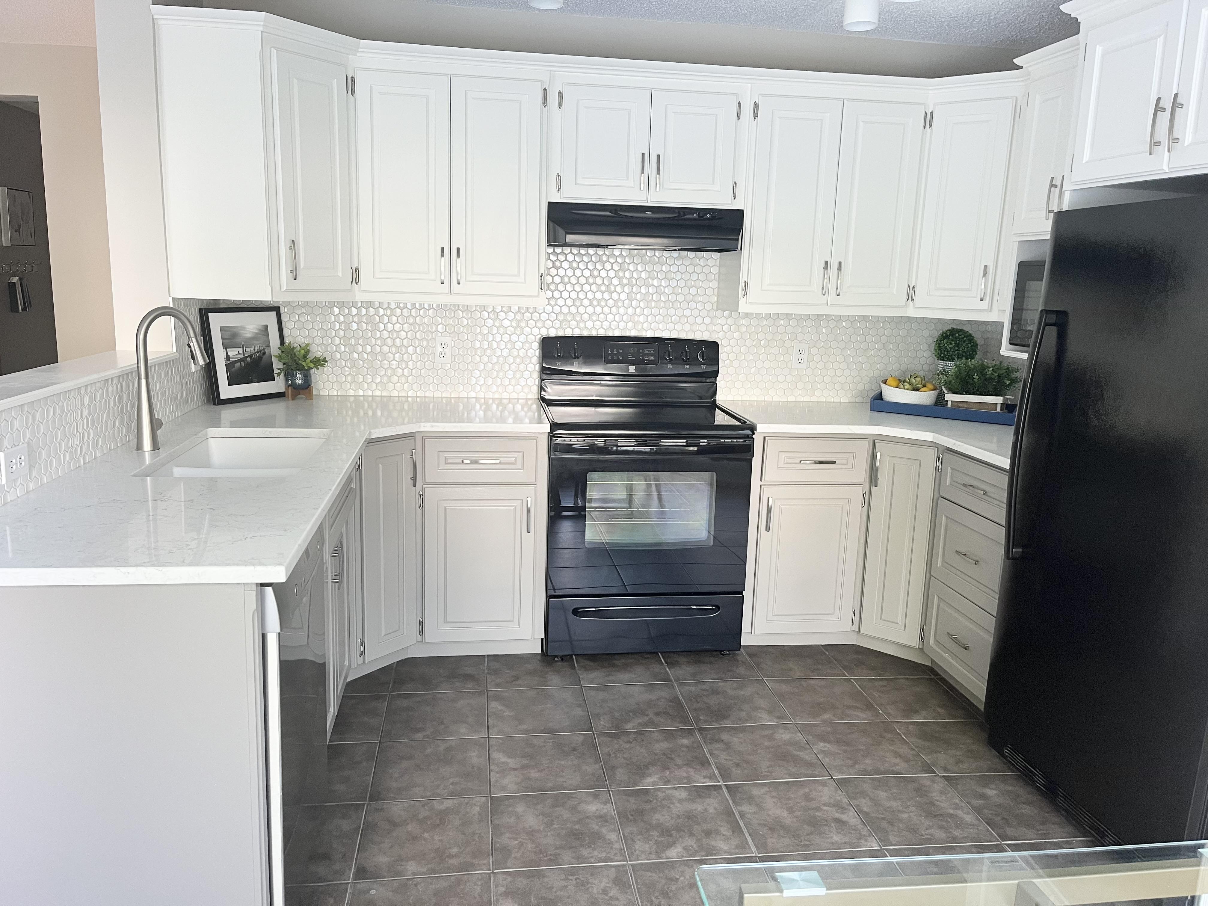 Staged kitchen with white cabinets, mosaic tile backsplash, black stove and range hood, white countertops, and dark tile flooring.
