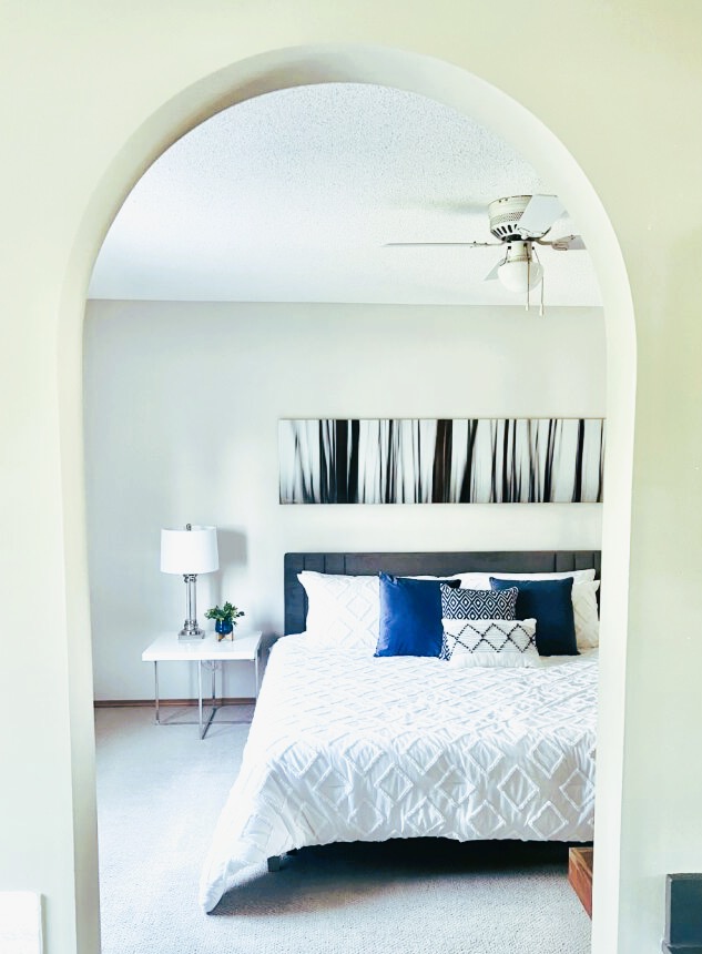 View through an arched doorway of a staged bedroom with white bedding, blue accent pillows, a gray headboard, and modern bedside table with lamp.