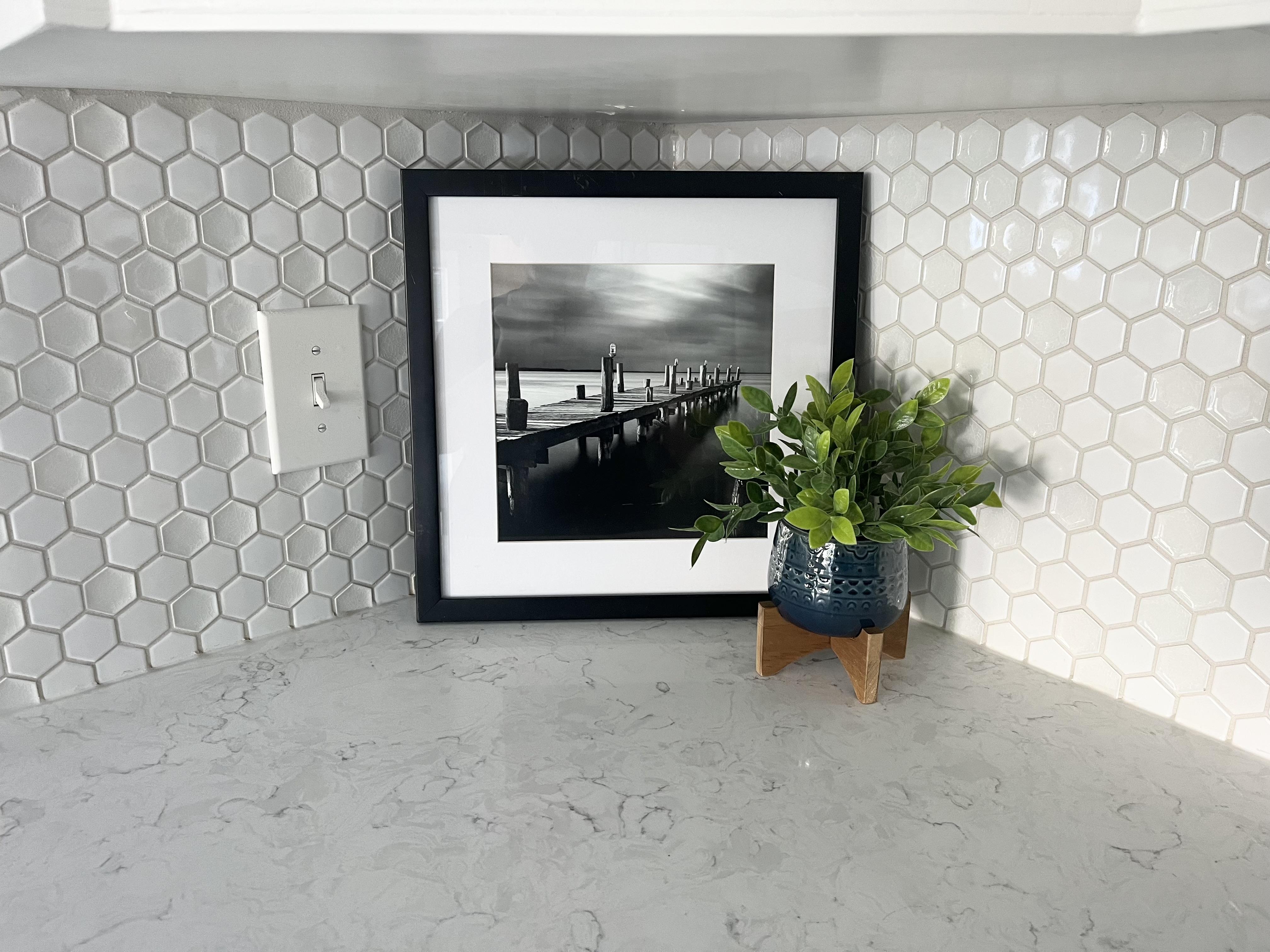 Kitchen countertop with a framed black-and-white photo and small potted plant styled against a white hex tile backsplash.