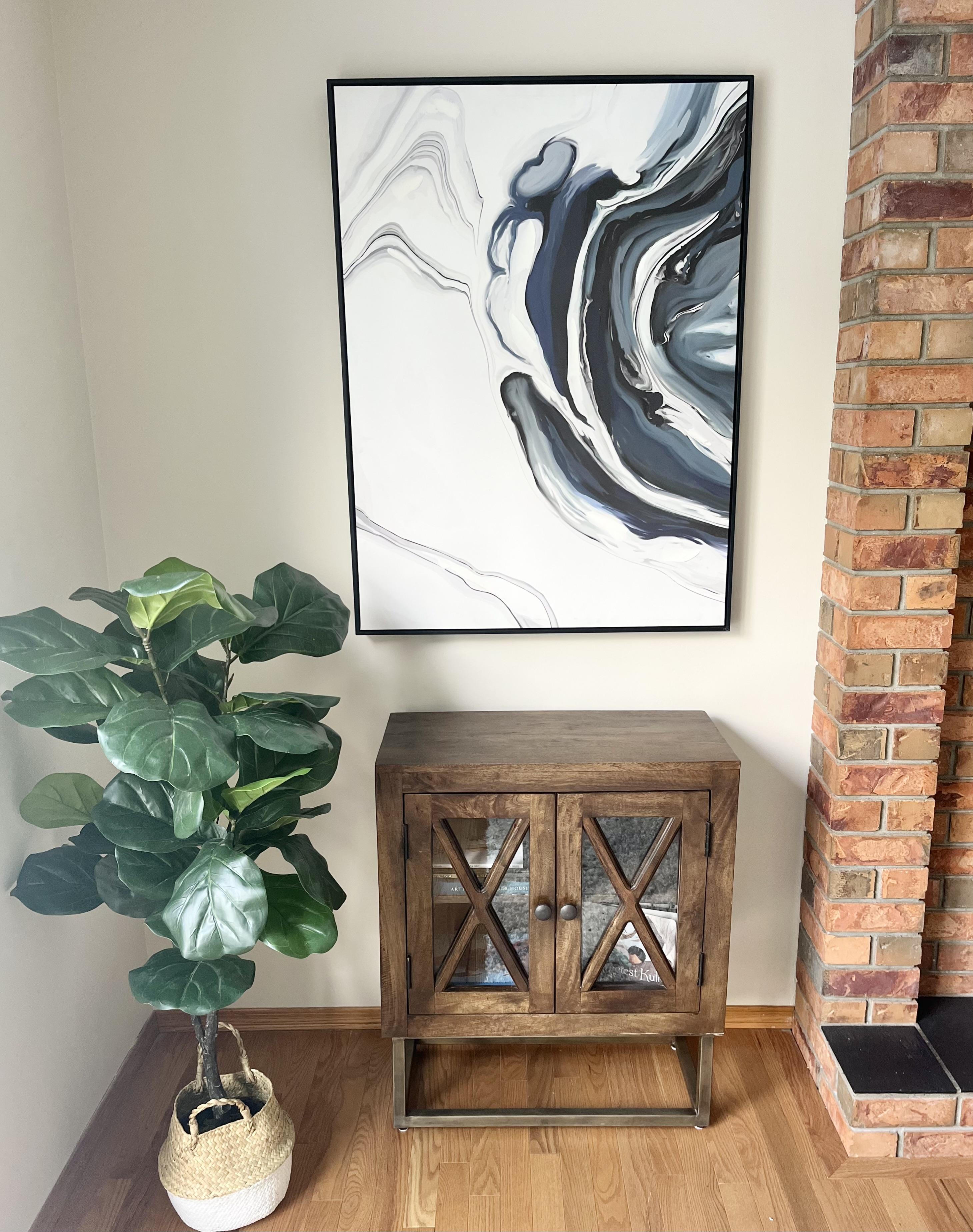 Decorative staging vignette with a wooden cabinet, large abstract wall art, and a leafy plant beside a brick fireplace.