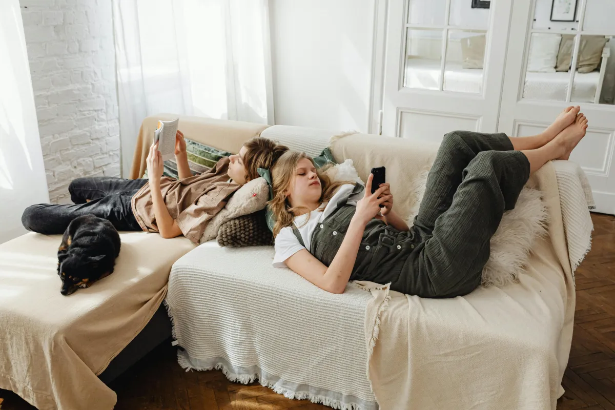 A girl, boy, and dog on a couch comfortable and reading their phones and books. 