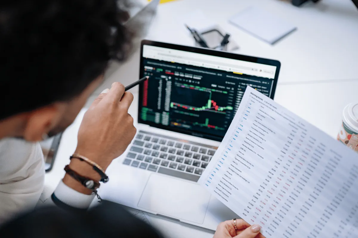 Person reviewing stock market data on a laptop while holding a printed report
