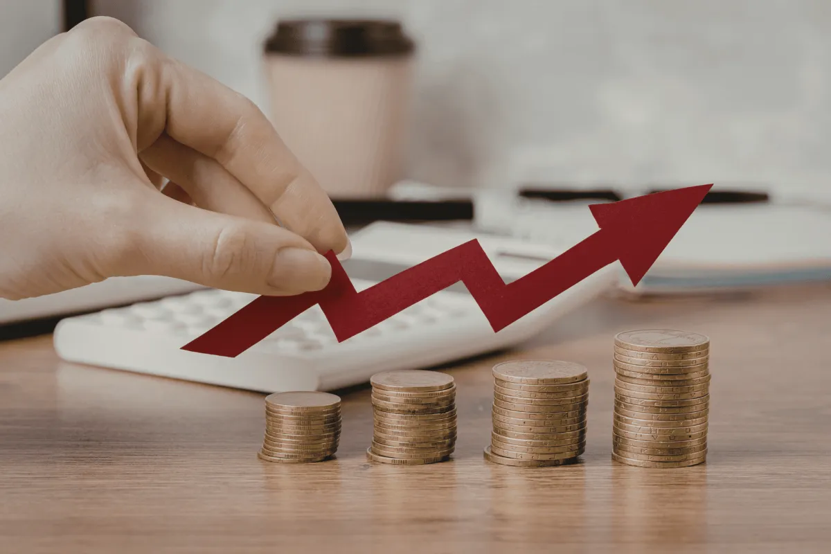 Hand holding red upward arrow above rising stacks of coins
