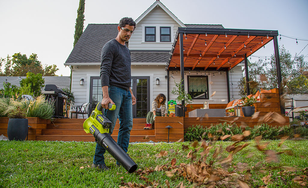 Homeowner using a leaf blower to clear yard debris near a deck and landscaped backyard in a suburban Mid-South home.