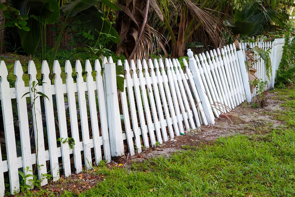 White wooden picket fence leaning and misaligned due to ground movement and aging fence posts.