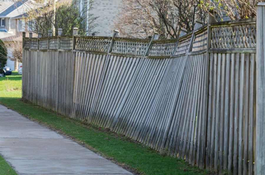 Tall wooden privacy fence severely leaning along a sidewalk due to failed posts and long-term weather exposure.