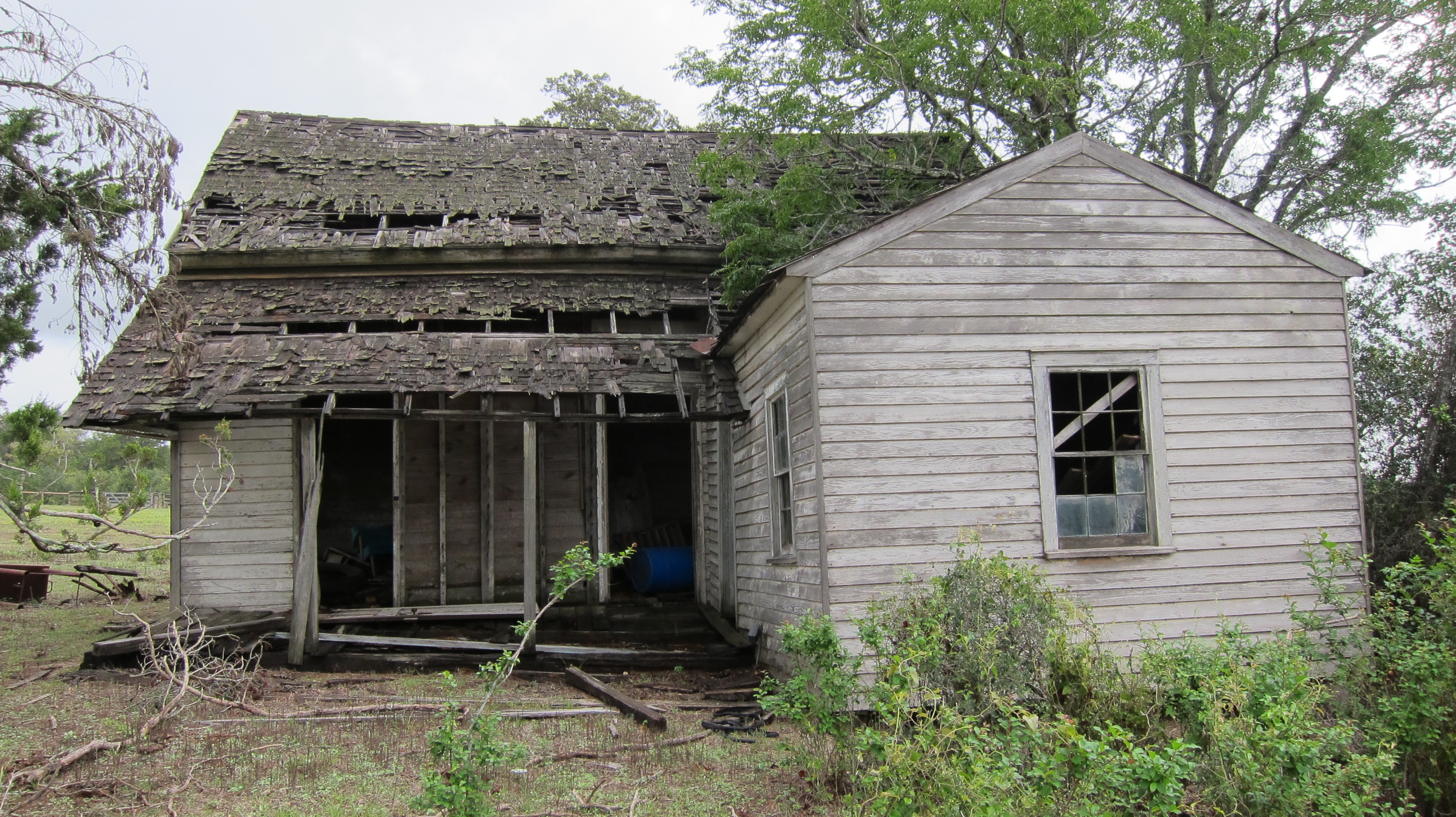 Severely deteriorated wooden house with collapsing roof, rotted siding, and exposed framing in a rural setting.
