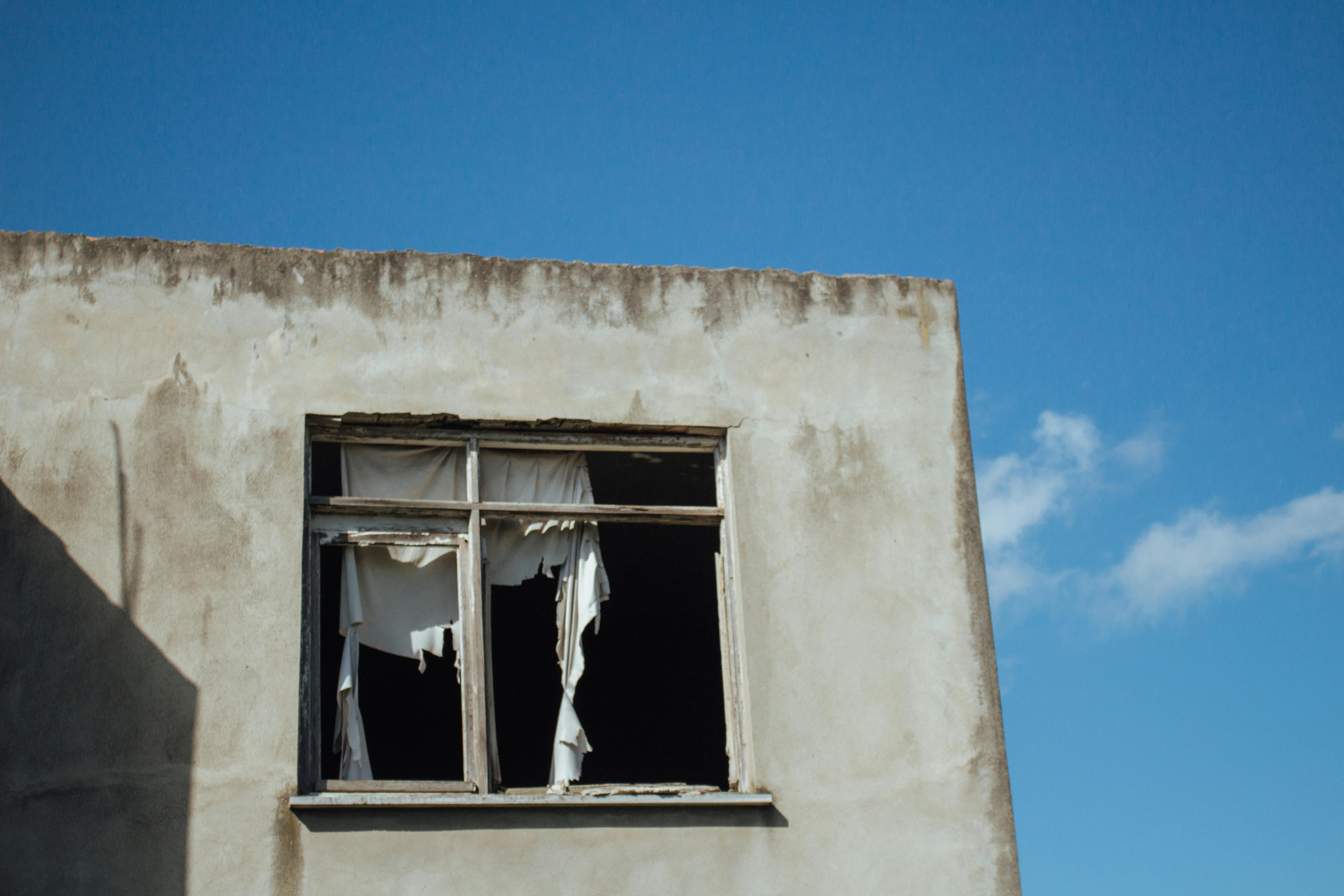 Broken window and cracked exterior wall on an aging building with peeling materials and exposed interior.
