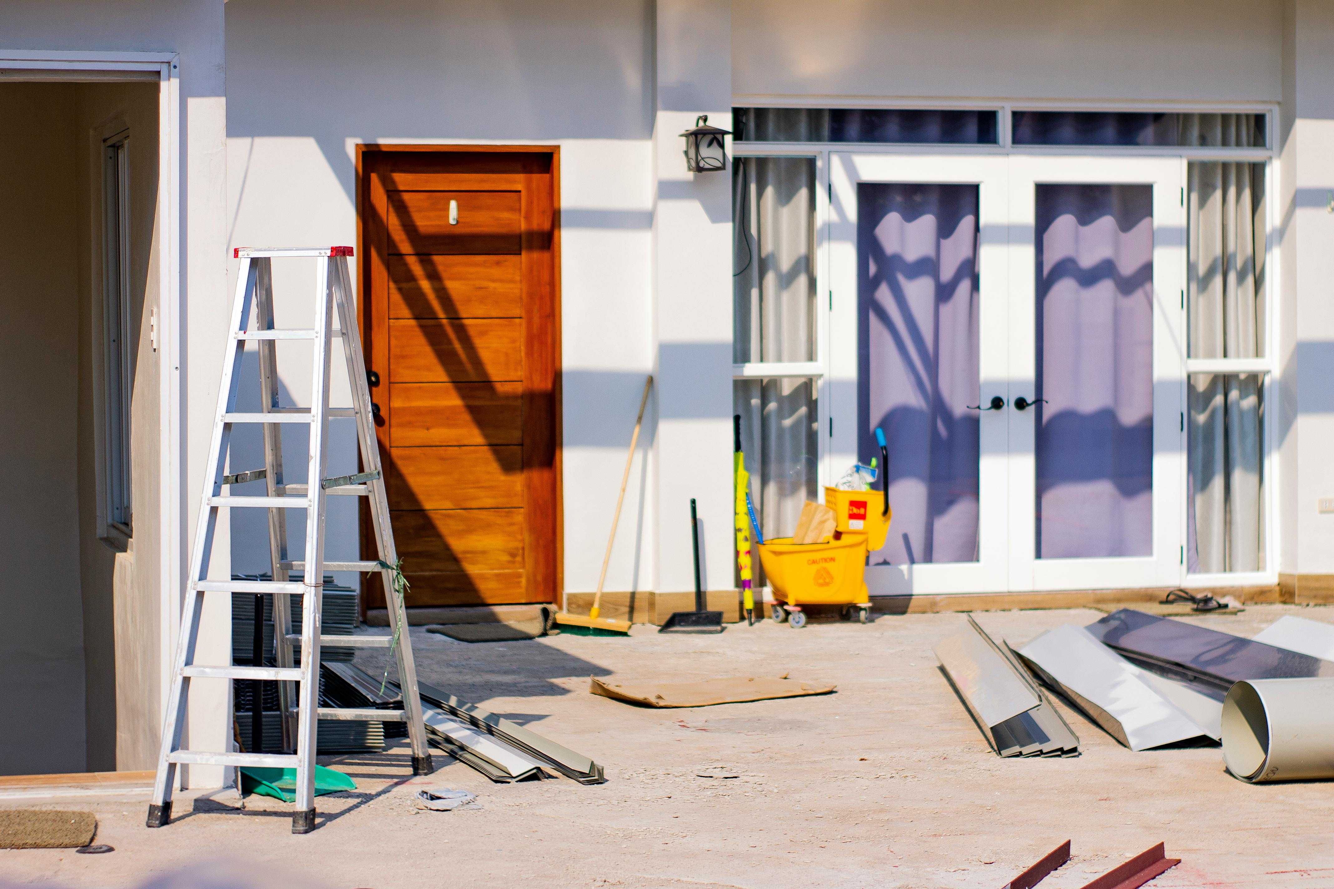 Exterior of a home during a repair project with ladder, tools, metal trim pieces, and materials staged on a concrete patio.