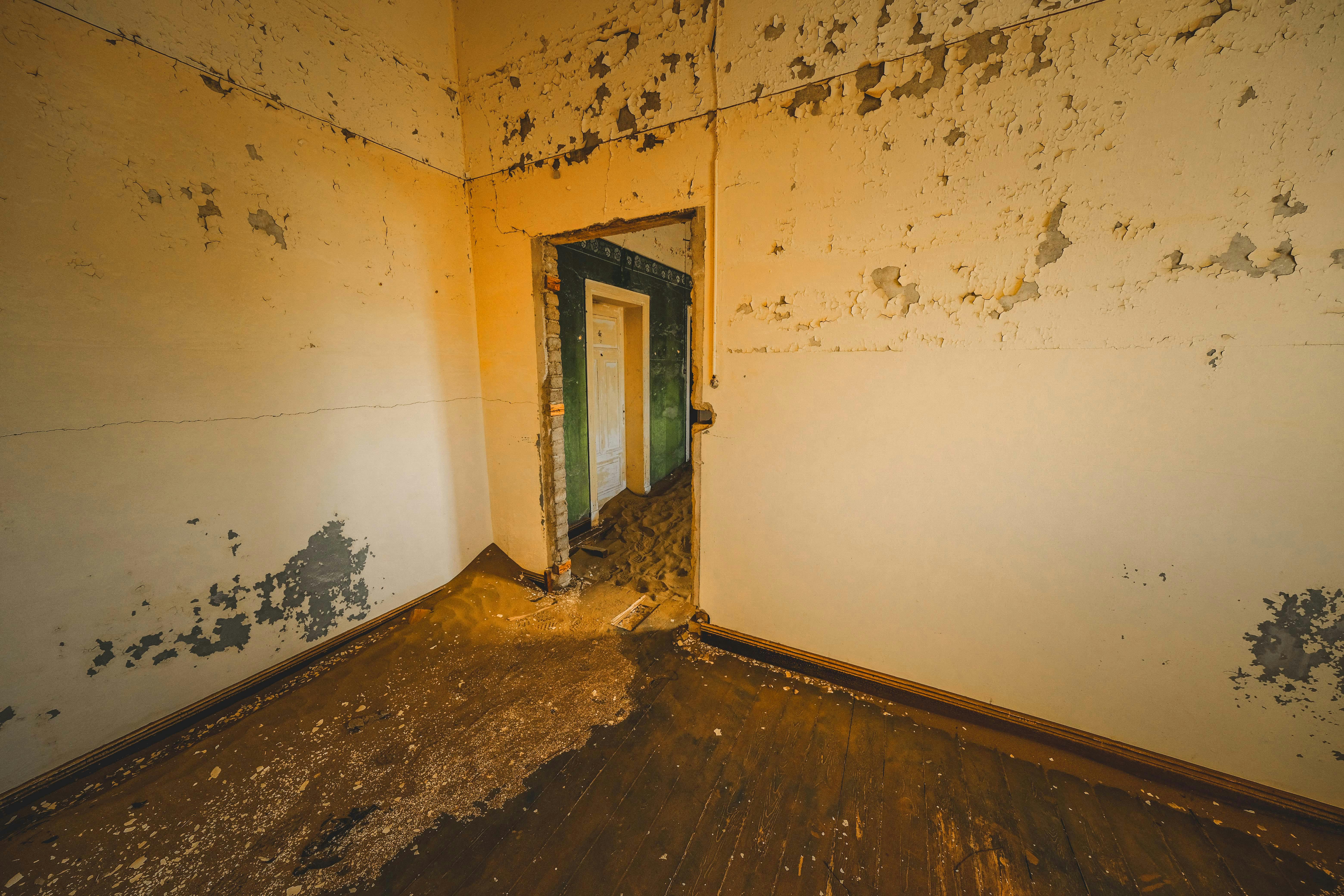 Empty interior room with peeling paint, wall cracks, and floor damage caused by water intrusion and long-term moisture exposure.