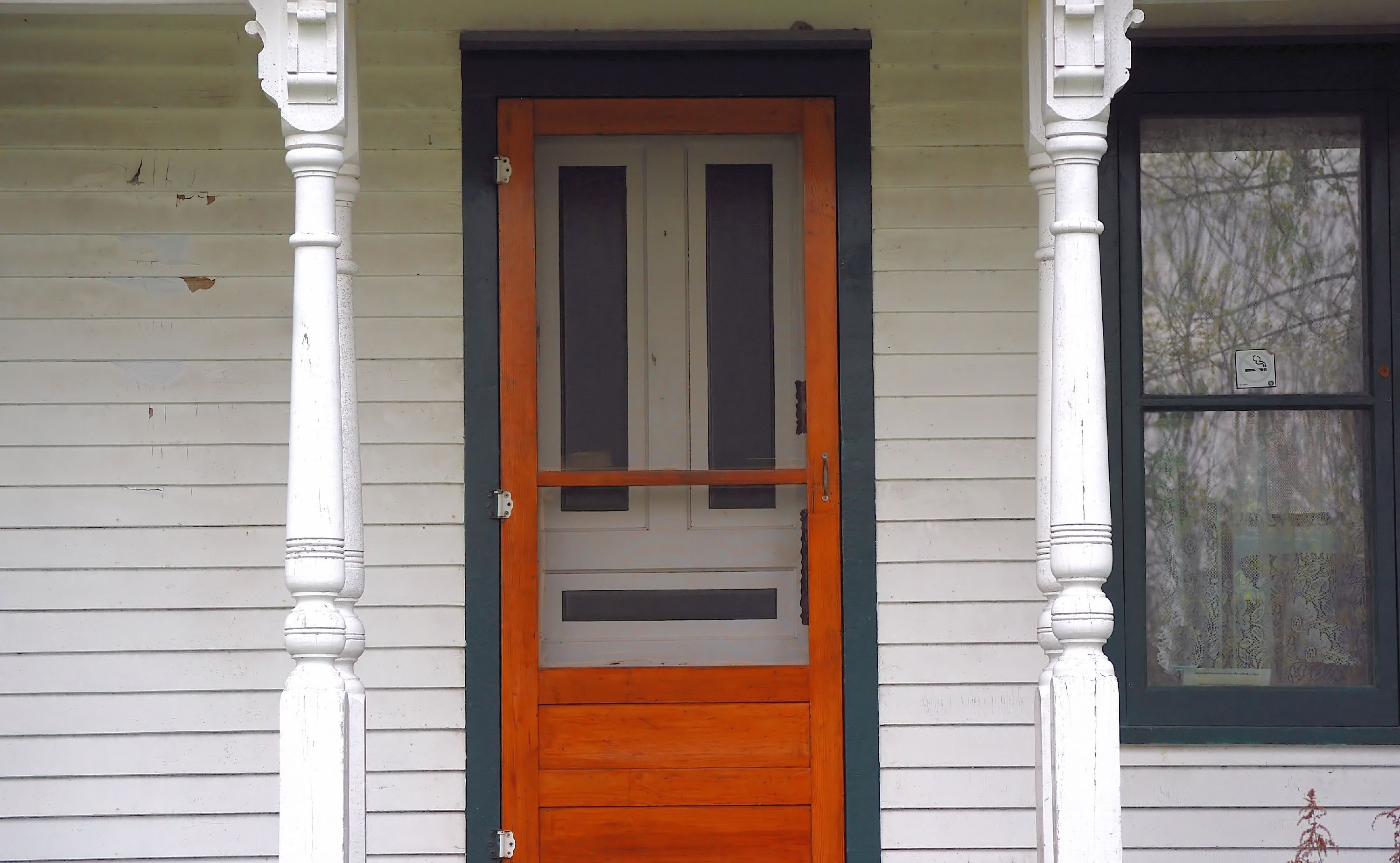 Older wooden screen door installed on a front porch with white siding, decorative columns, and adjacent window.