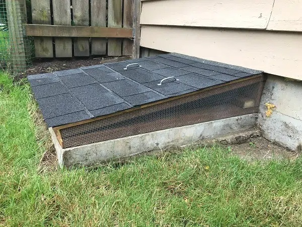 Exterior crawl space vent opening with protective screened cover and shingled access panel installed at the base of a home foundation.