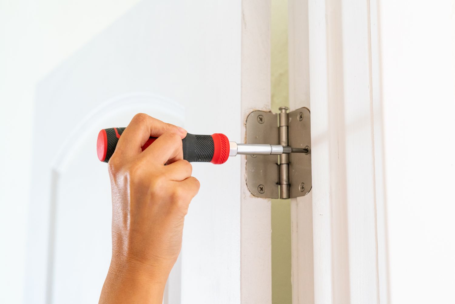 Hand using a screwdriver to adjust door hinges on an interior door to fix alignment and sticking issues.