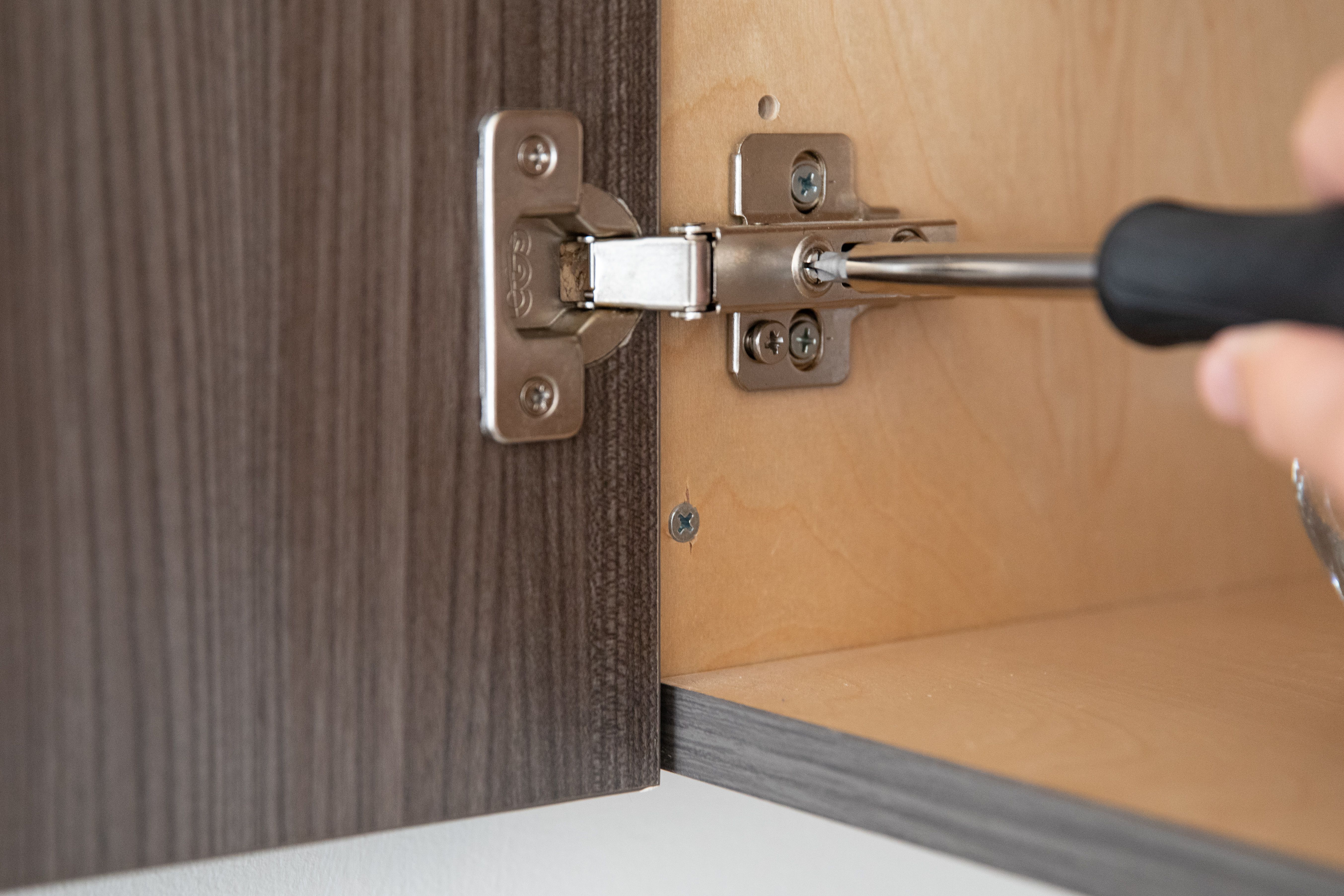 Close-up of a cabinet hinge being adjusted with a screwdriver inside a kitchen cabinet.