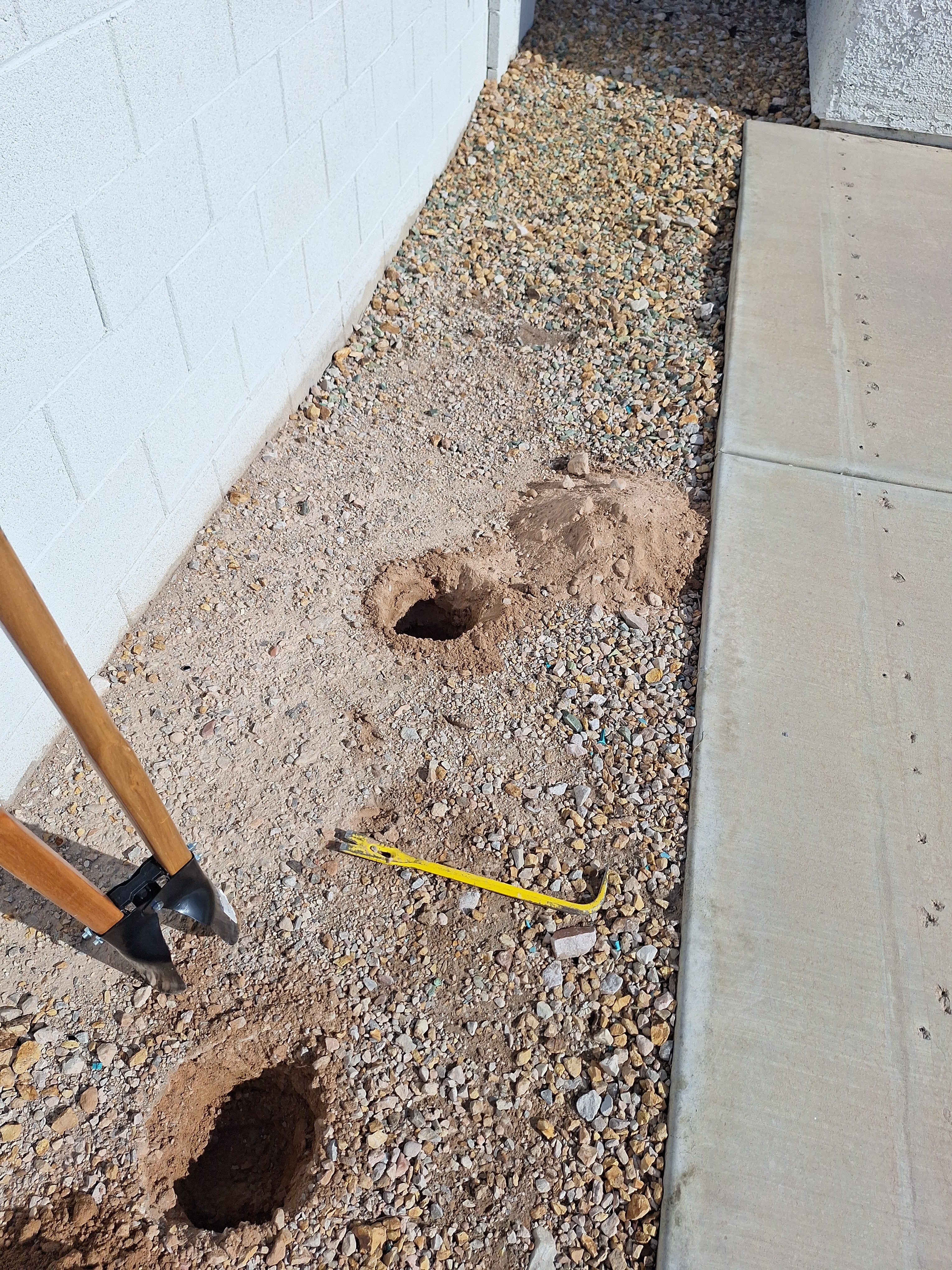 Two freshly dug holes for fence posts are visible next to a white block wall and concrete path in a Las Vegas yard, with a post-hole digger and pry bar, indicating fence repair.