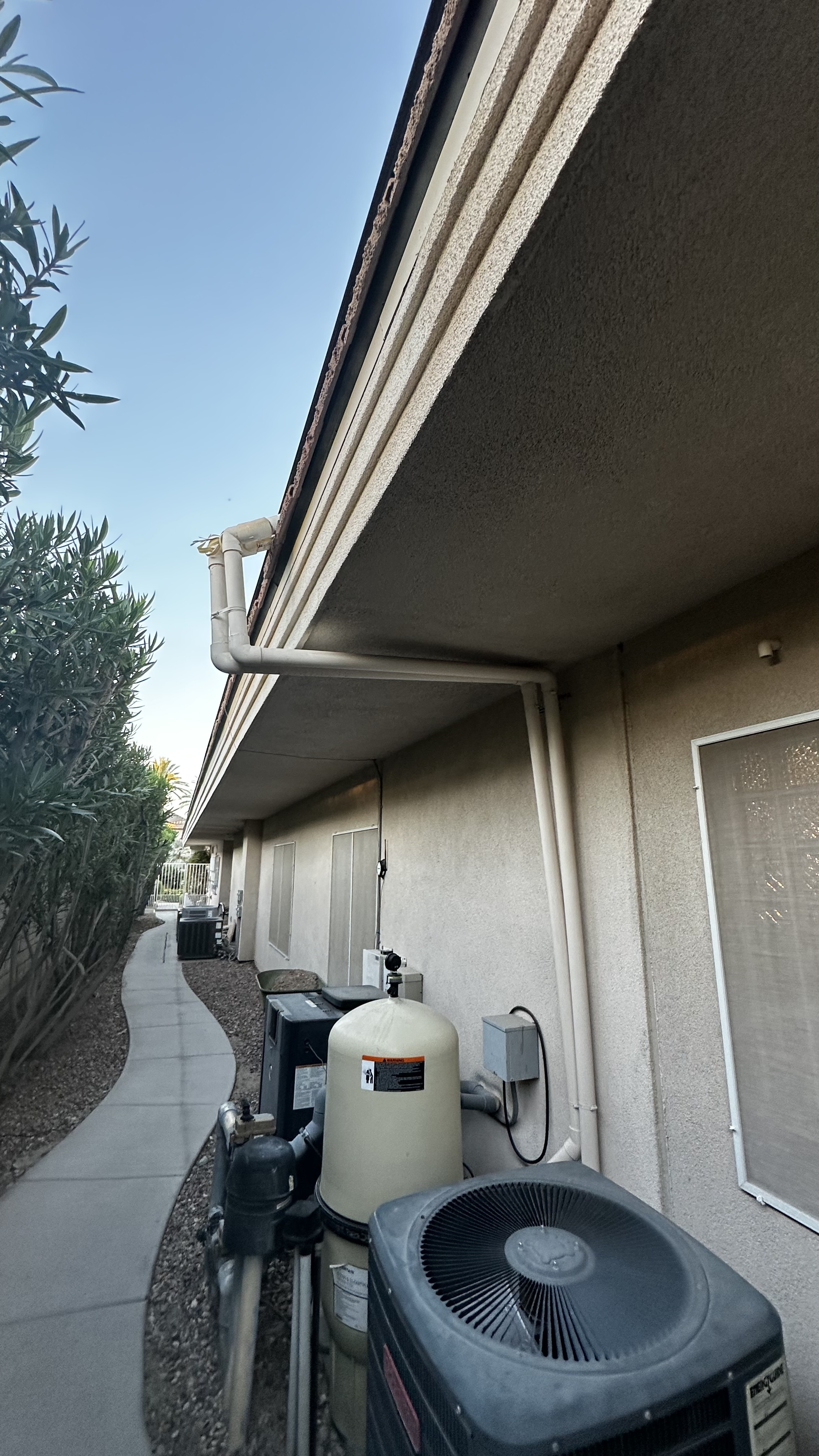 A view of a residential home's roof edge showing gutters and downspout, with a pool filter and air conditioning unit below, and a walkway and landscaping to the side