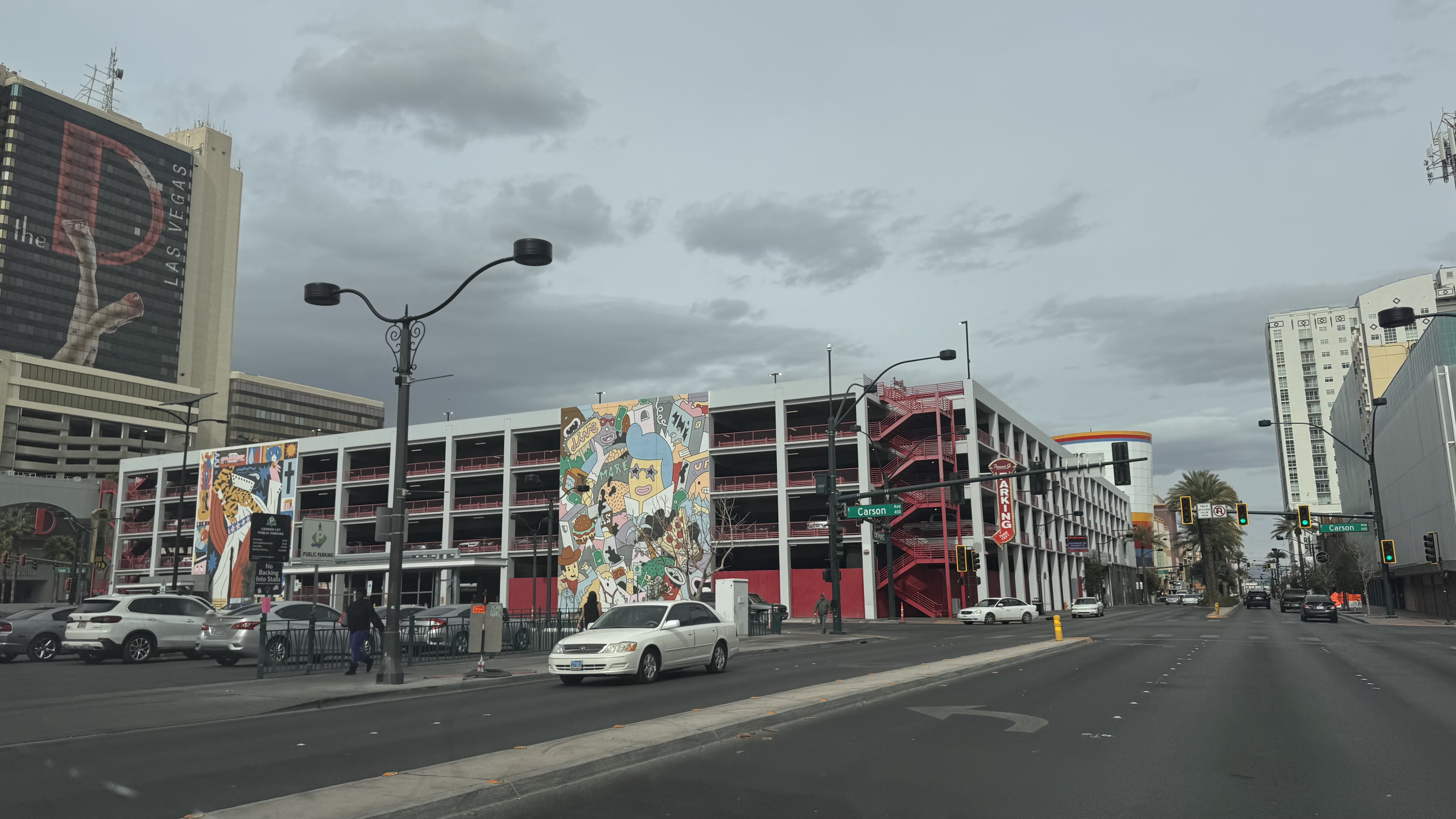 Urban Las Vegas street scene showing Carson Avenue intersection with a multi-story parking garage featuring colorful murals, street lamps, and vehicles under overcast skies.
