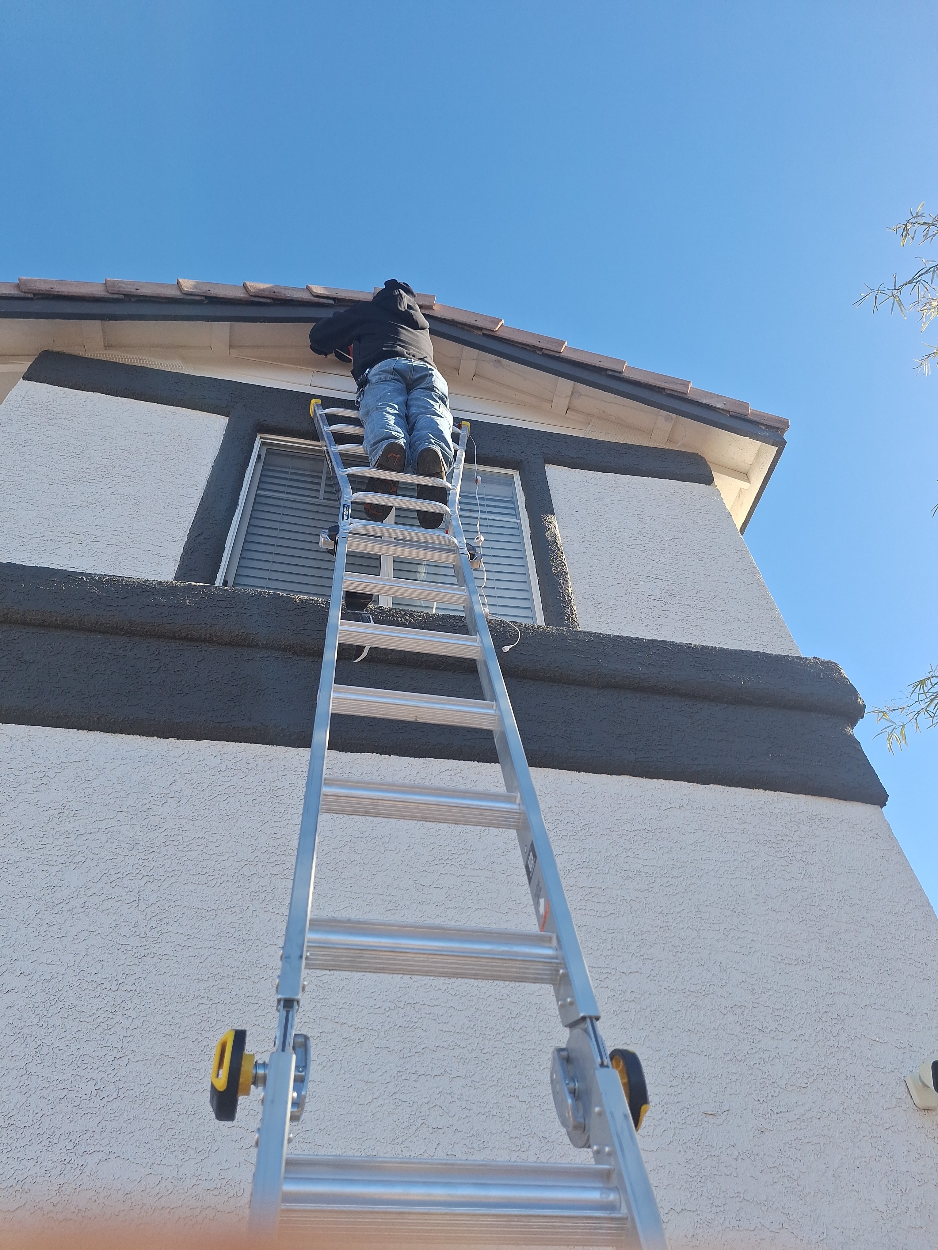 A handyman on an aluminum ladder repairs the upper section of a stucco house with dark trim, focusing on the roofline and window area under a clear blue sky.
