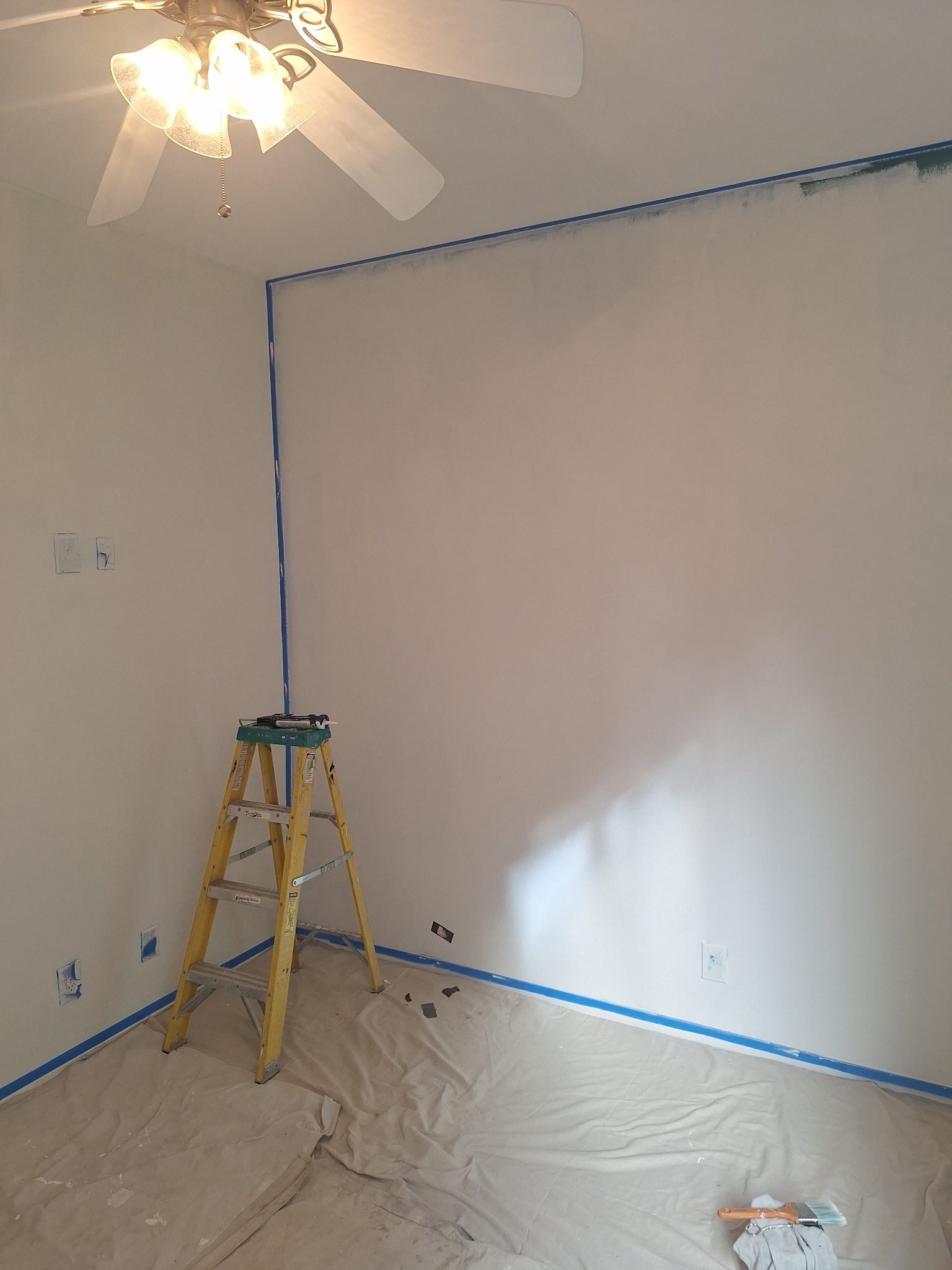 A room with white walls being prepared for painting, showing blue painter's tape along edges, a yellow ladder, drop cloths on the floor, and a ceiling fan with lights. This depicts the work of a Las Vegas handyman for rental property turnover.