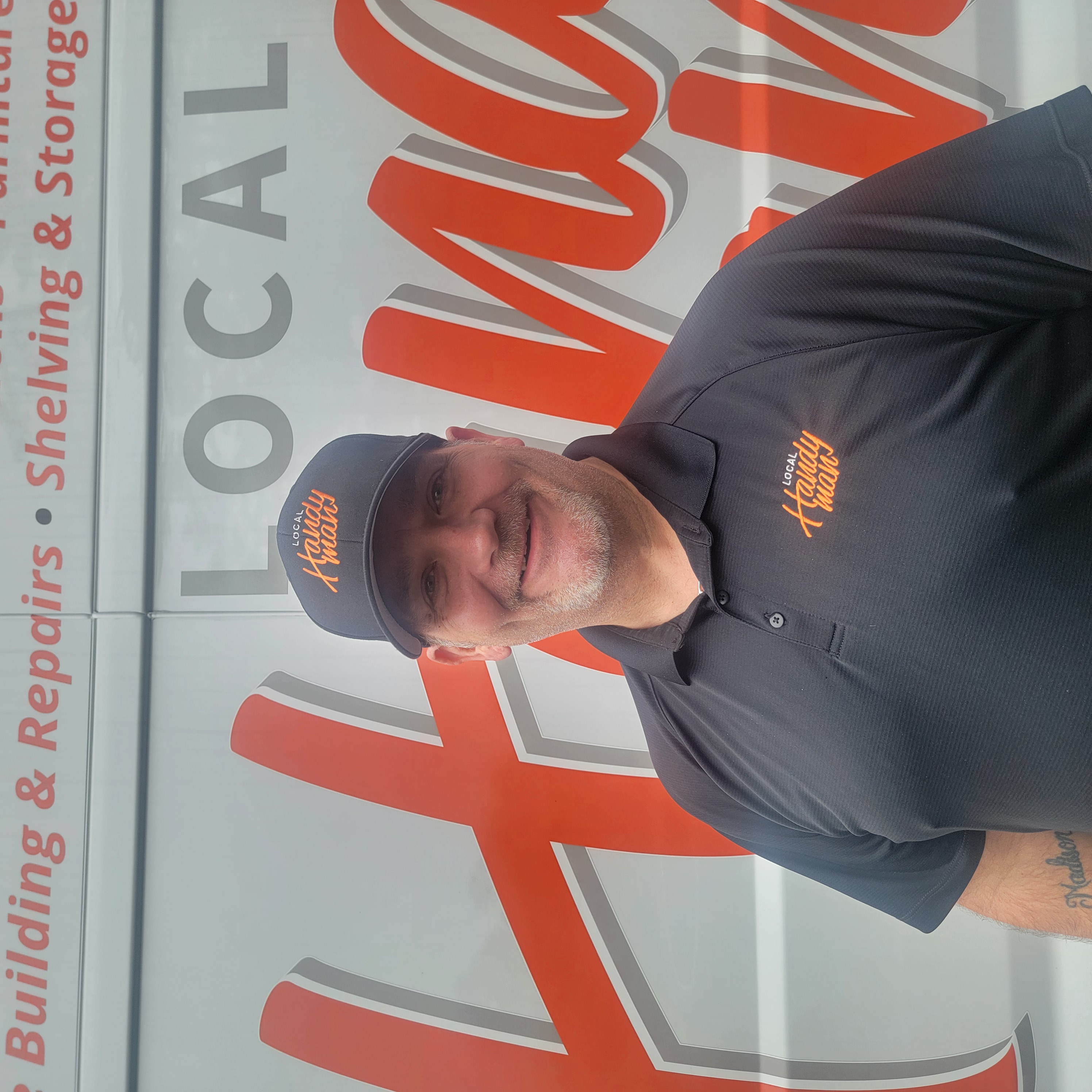 Tony Howell wearing a Local Handyman cap and polo, posing in front of a company van.