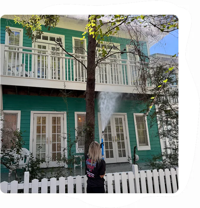 Person pressure washing the second-story exterior of a green house with white trim and picket fence.