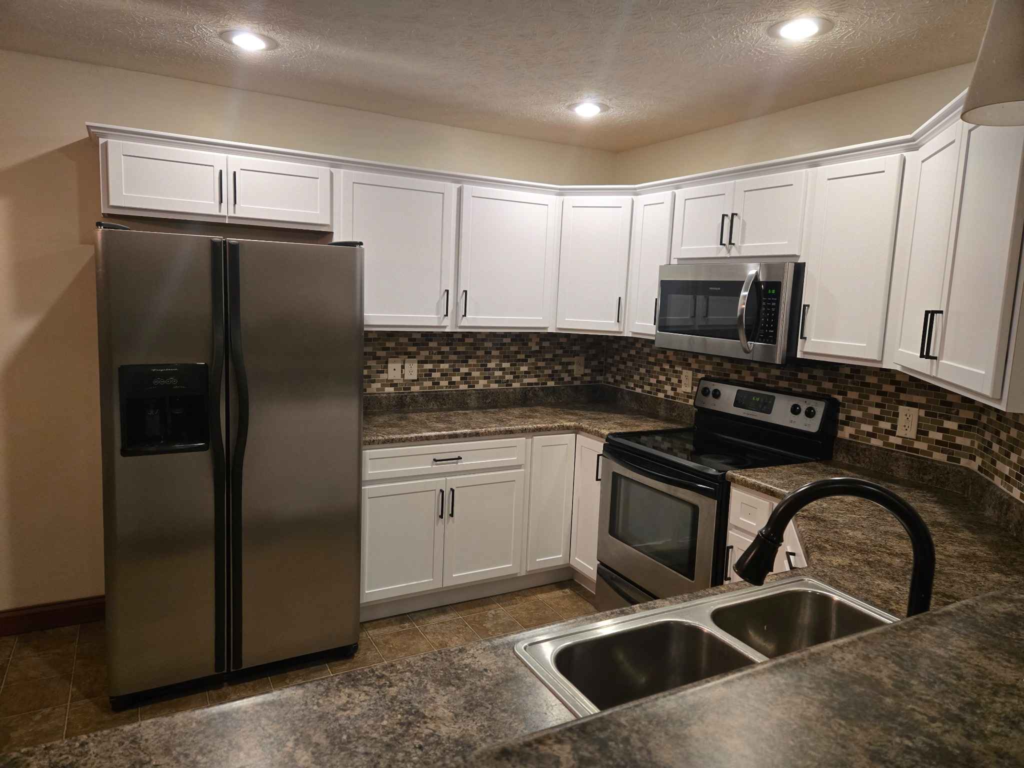 Kitchen with white shaker cabinets, stainless steel appliances, mosaic tile backsplash, and brown stone-look countertops under recessed lighting.
