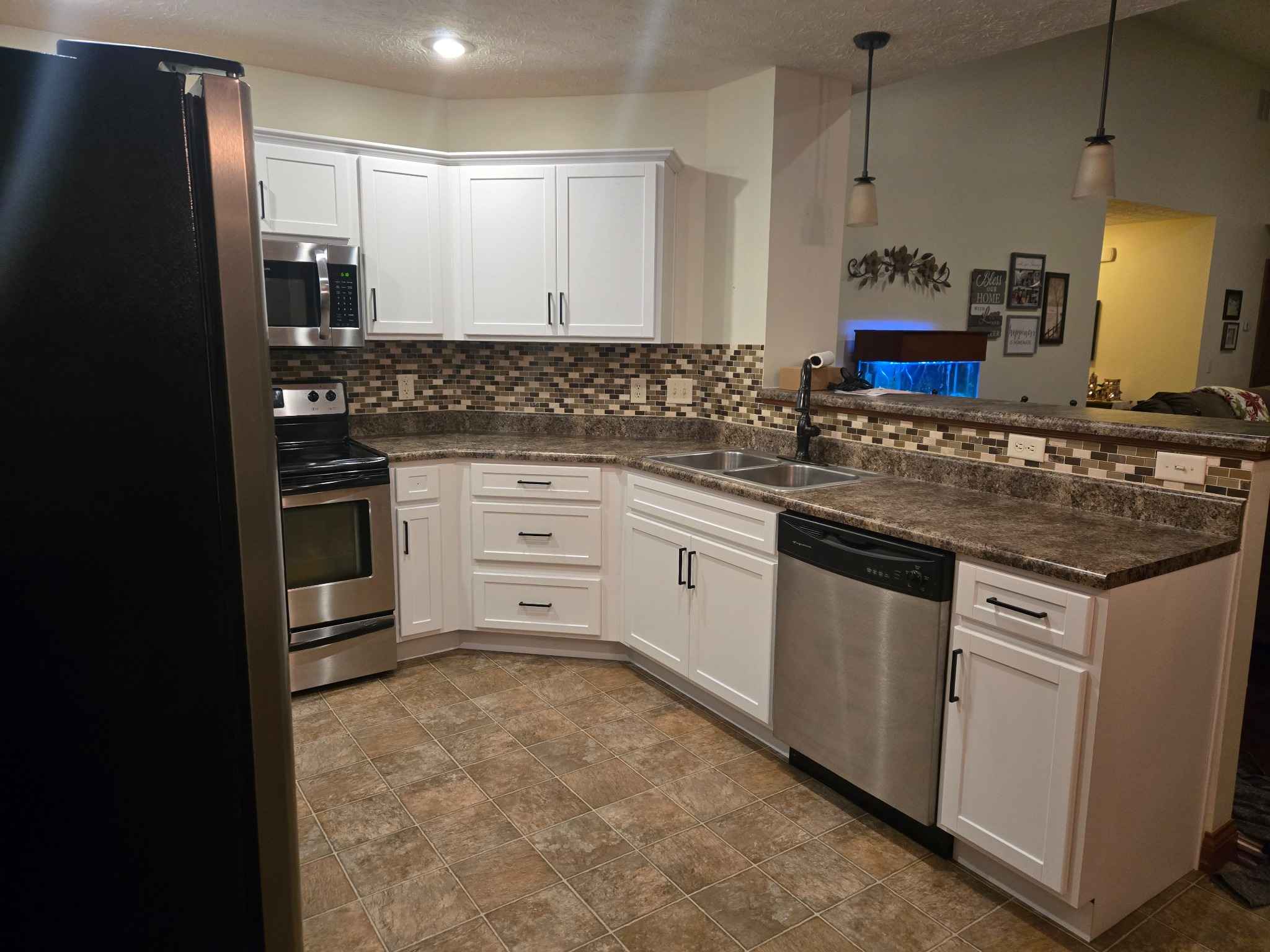 Kitchen with white shaker cabinets, stainless steel appliances, mosaic backsplash, double-basin sink, and brown stone-look countertops viewed from the peninsula area.