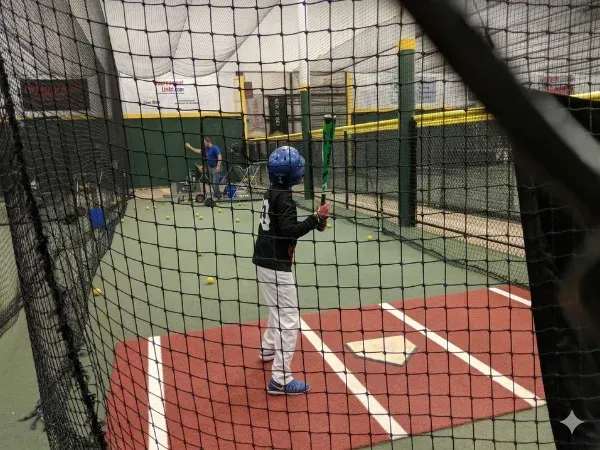 A young boy in a blue helmet and white uniform stands in an indoor batting cage holding a lime green bat.
