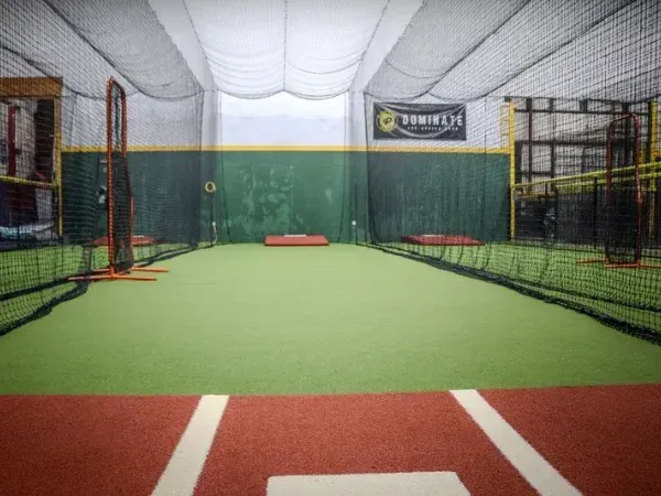 An empty indoor baseball batting cage with green turf, dark green walls, and red clay-colored base path.