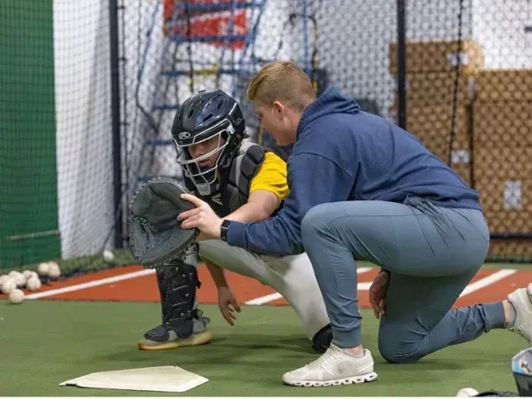A baseball coach kneels beside a young catcher in full gear, demonstrating a catching technique indoors.