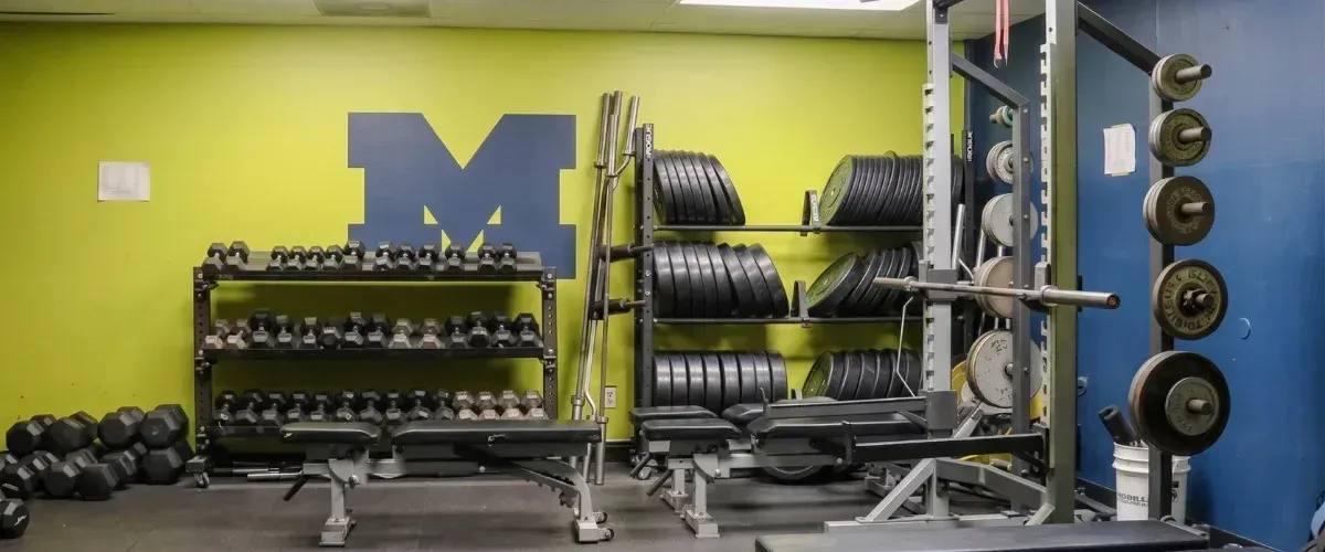 A wide view of a gym weight room with shelves of dumbbells, barbells, and weights, a blue M logo on the lime green wall.