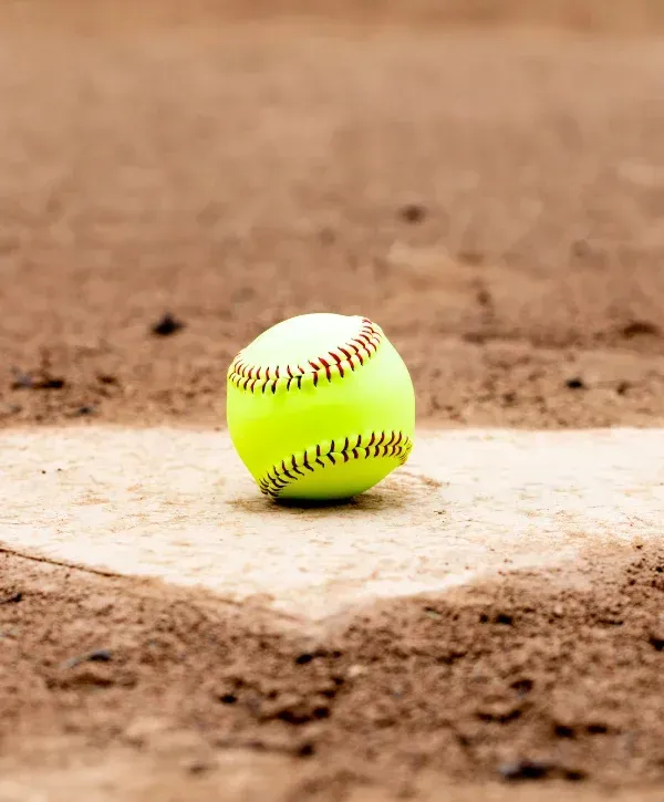 A bright yellow softball with red stitching rests on a white foul line within a brown dirt baseball field.