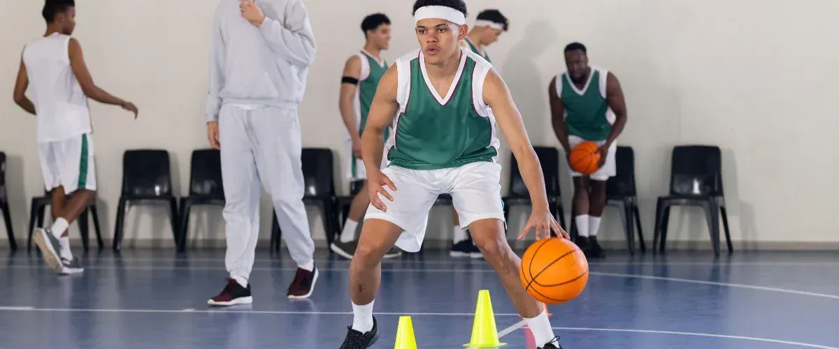 A young basketball player in black and red shorts dribbles the ball on a wooden court during practice.