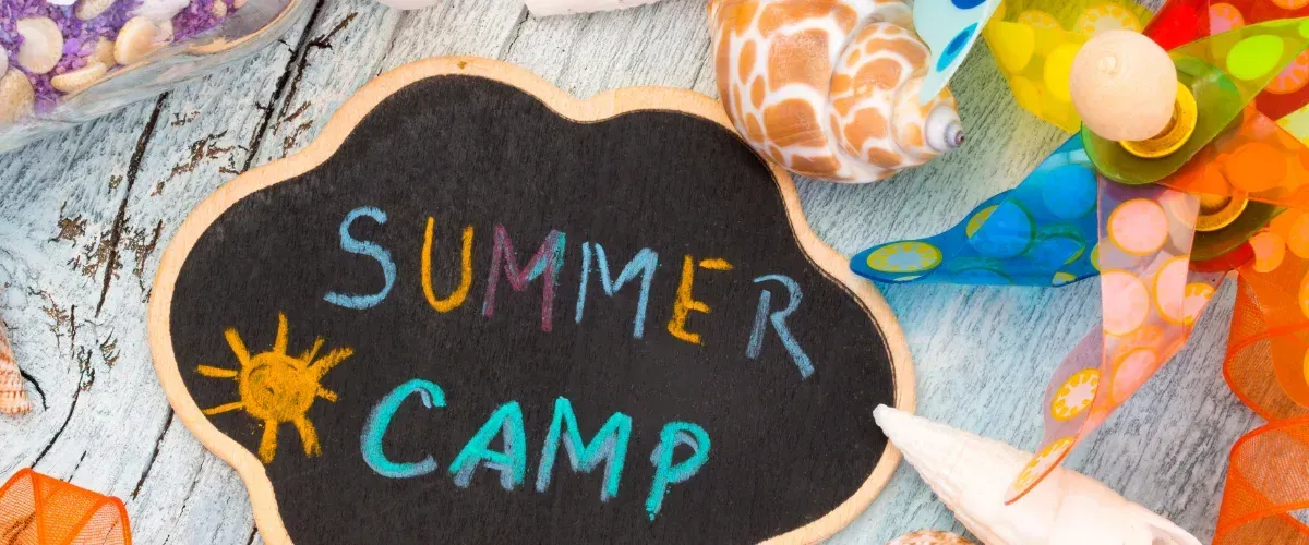 A chalkboard sign saying "SUMMER CAMP" with a sun drawing, surrounded by a pinwheel, shells, and colorful ribbons.