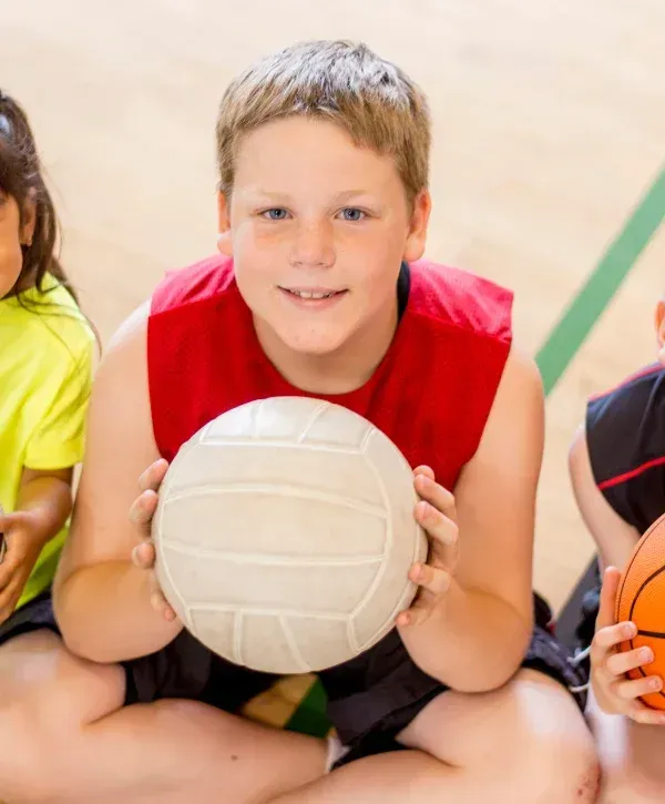 A smiling boy with freckles and blue eyes holds a white volleyball, sitting on a gym floor with other children.