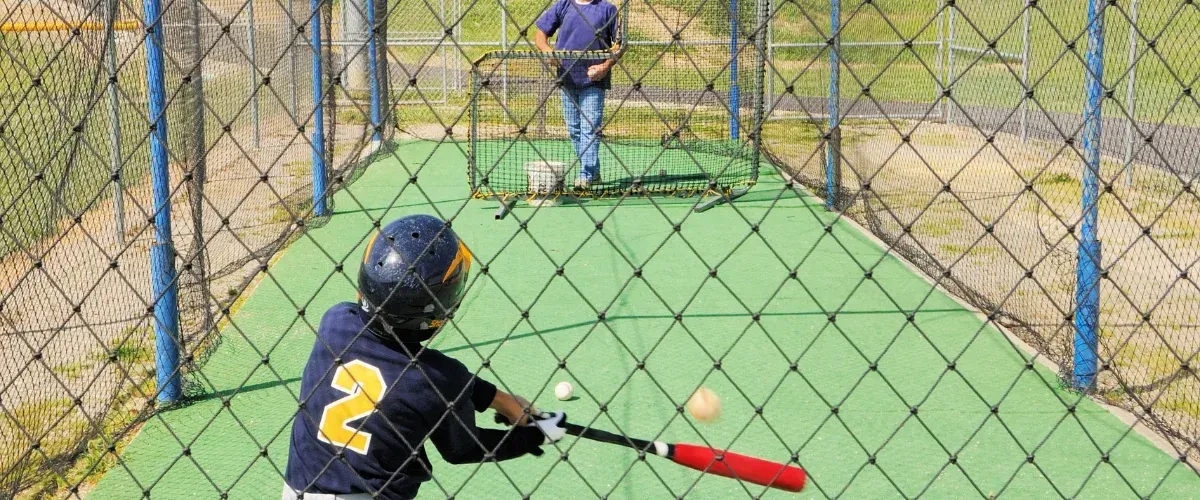 A young boy in a blue shirt and shorts takes a powerful swing at a yellow baseball at a batting cage.