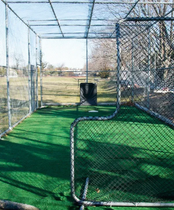 An empty outdoor batting cage with green turf, a white home plate, and a black pitching screen, surrounded by chain link fences on a sunny day.