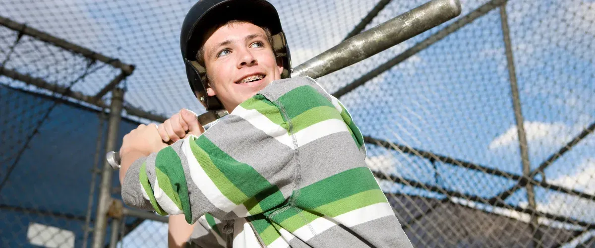 A smiling young male baseball player with braces and a helmet, holding a bat, looking upwards with an upbeat expression.