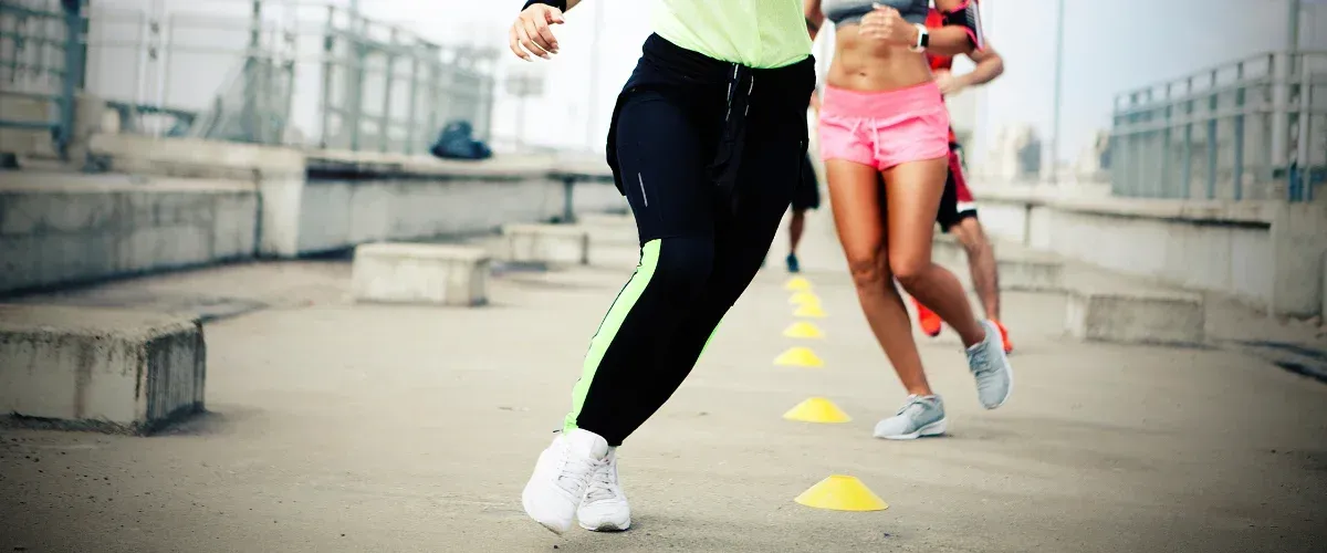 Close-up of athletes' legs and torsos as they run an agility drill with yellow cones on a concrete surface.