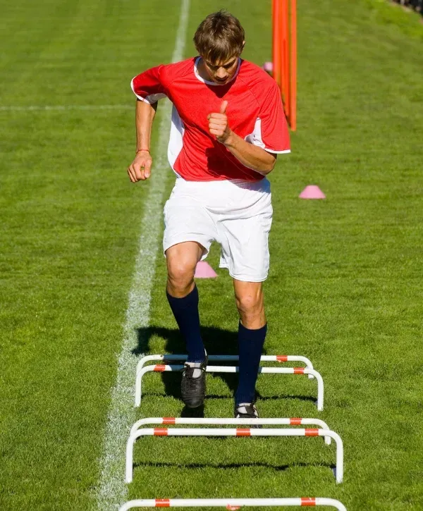 A young male soccer player in a red and white uniform performs agility drills over small hurdles on a green field.