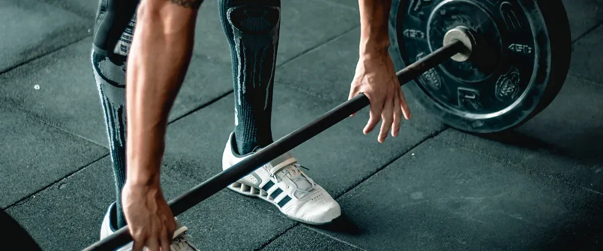 An athlete with muscular arms and calves wearing white training shoes prepares to deadlift a barbell with heavy weights.