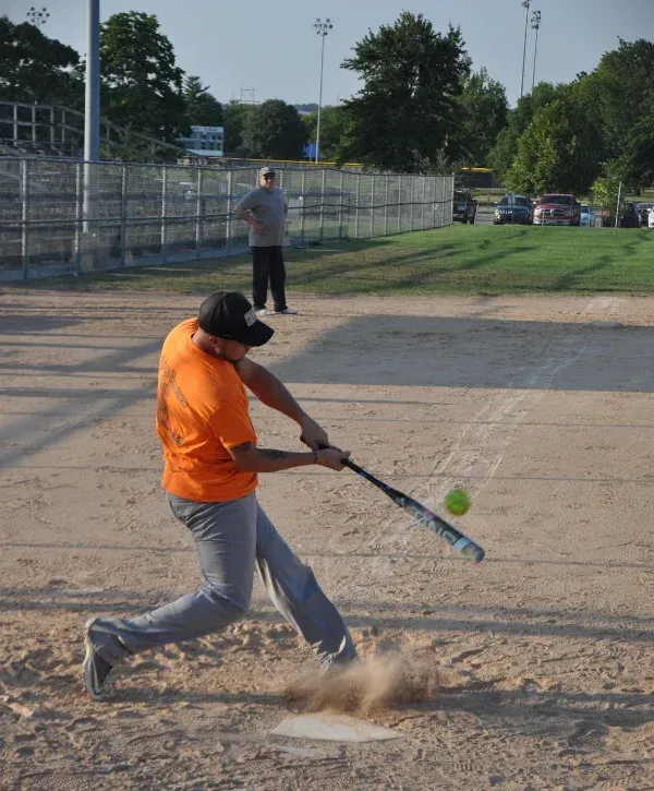 A female softball player in a black helmet and green/white jersey swings a colorful bat at a yellow ball on a dirt field.