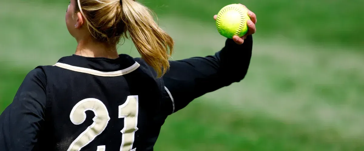 A female softball player in a black helmet and green/white jersey swings a colorful bat at a yellow ball on a dirt field.