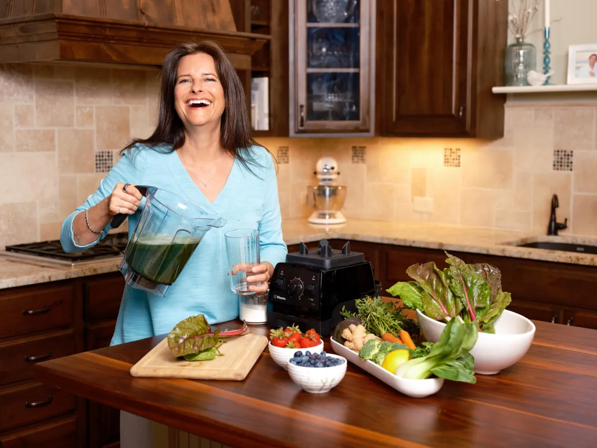 Shellee Methe RN smiling in kitchen making a green smoothie with fresh produce, holistic wellness and faith-based weight loss coach