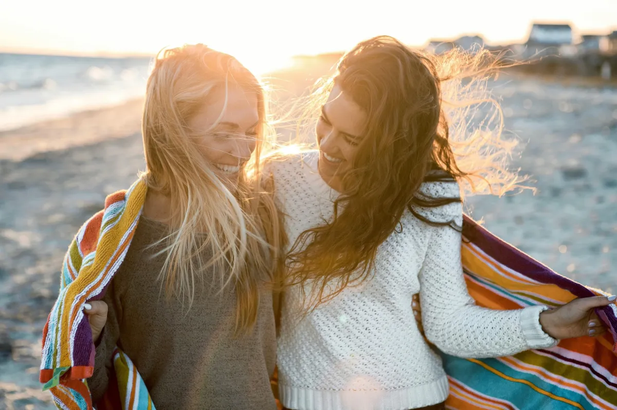 Two women smiling and wrapped in a colorful blanket on a sunny beach, enjoying the breeze and each other's company.