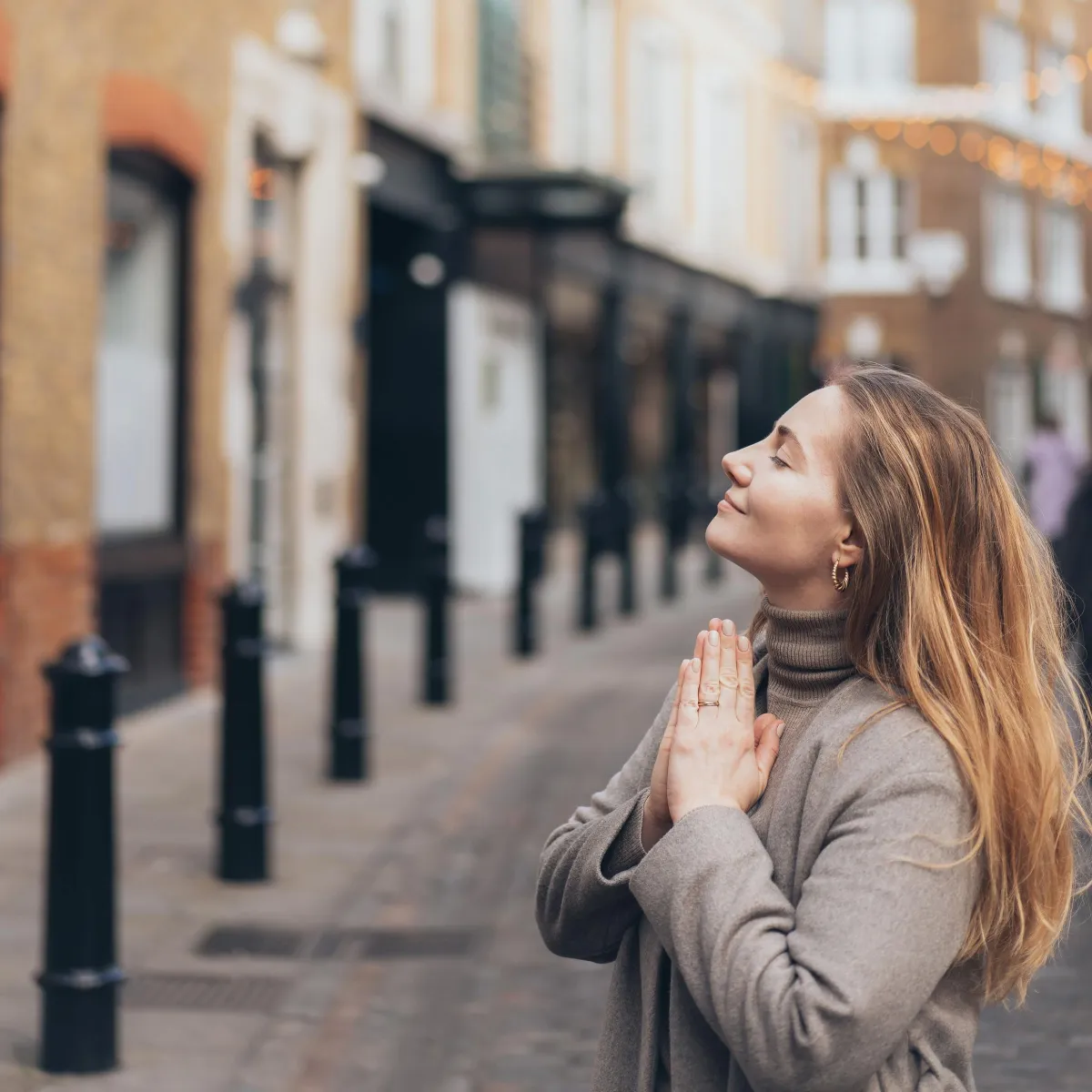 A serene woman with blonde hair practices gratitude on a charming city street, eyes closed and hands in prayer position, embodying peace and mindfulness.