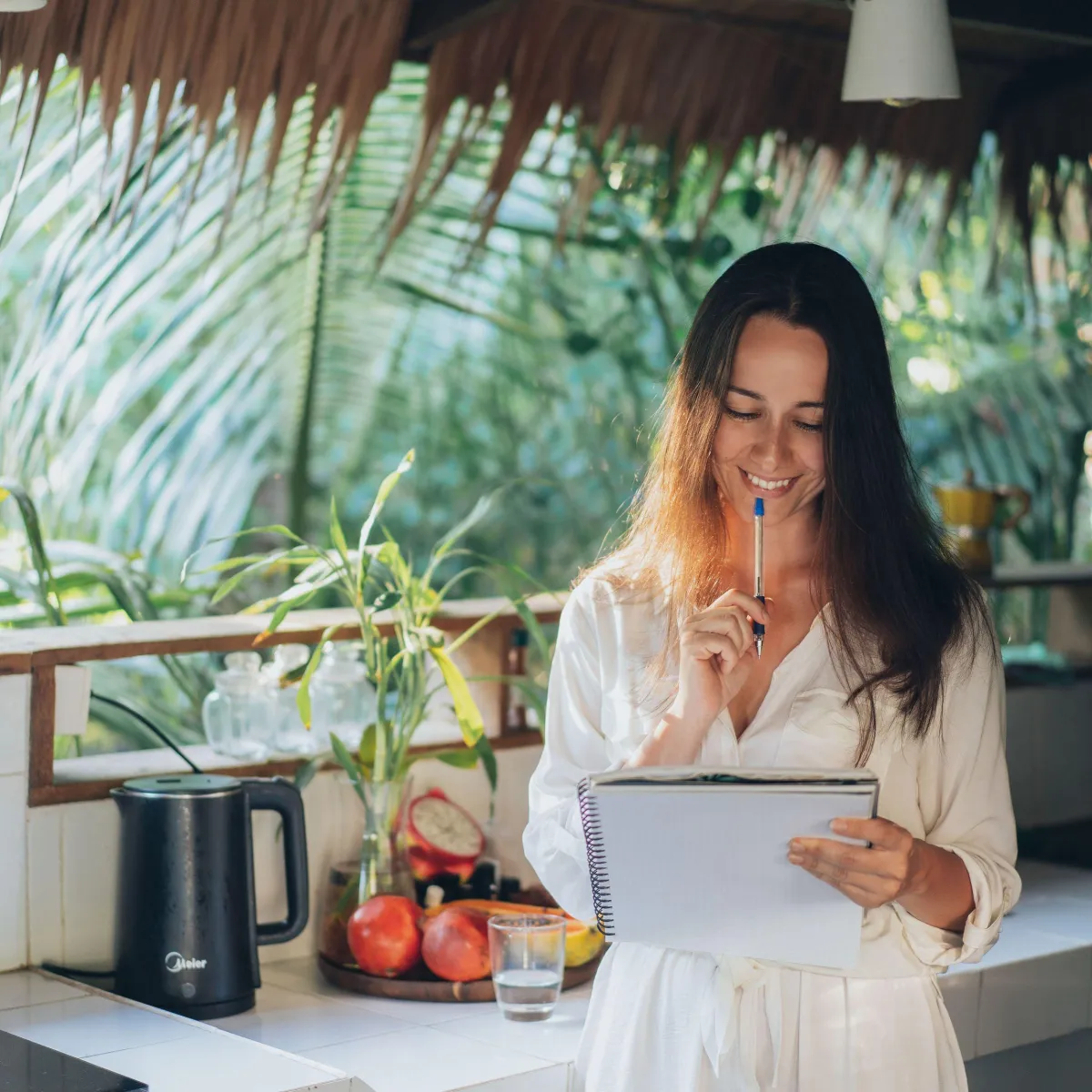 A woman smiles while writing in a notebook, enjoying a peaceful moment in a tropical, sunlit kitchen. She embodies mindfulness and creativity, surrounded by lush greenery and fresh fruits, reflecting a balanced and fulfilling lifestyle.